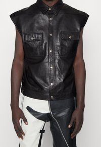 Svart läder ärmlös skjorta med två framfickor, metallisk tryckknappar och kontrasterande sömmar. Tyget har en slät, glänsande yta.