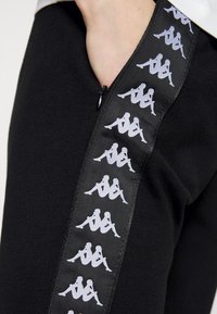 Svarta joggers med en dragkedjeficka och sidopanel med upprepade vita logomönster, tillverkade av ett mjukt, stretchigt tyg.