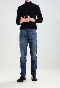 Suéter negro de cuello alto combinado con jeans de mezclilla azules de corte relajado, con un sutil desgaste, y zapatos de charol oscuros.