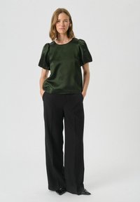 Top verde in satin con maniche corte a sbuffo, abbinato a pantaloni neri a gamba larga. L'outfit presenta una texture liscia e un design minimalista.