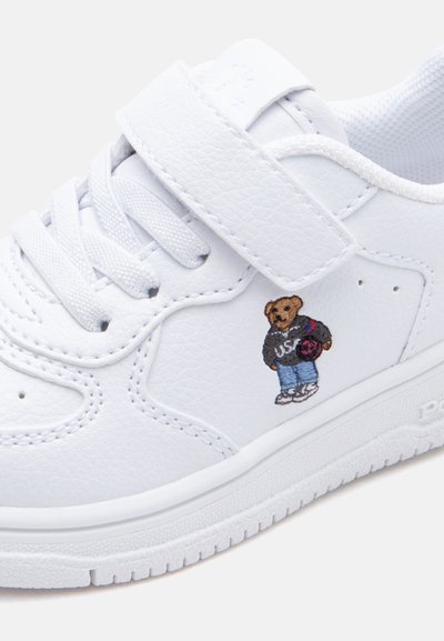 Λευκό δερμάτινο αθλητικό παπούτσι με κορδόνια και ιμάντα βέλκρο, με μικρή κεντημένη αρκουδίτσα που φοράει φούτερ USA και τζιν.