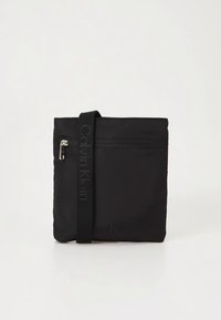 Čierna taška z nylónu so zipsovým vreckom, širokým textilným popruhom a reliéfne vyhotoveným logom. Obdĺžnikový tvar s hladkým povrchom.