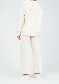 Světle béžová oversize mikina s žebrovanými manžetami, ve spojení s odpovídajícími širokými kalhotami a bílými slip-on botami, na jednobarevném pozadí.