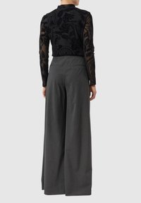 Kobieta ubrana w szare, szerokie spodnie z wysokim stanem i czarną, transparentną bluzkę z długim rękawem i kwiatowym wzorem, zwrócona tyłem.