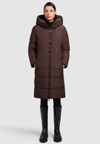 Manteau rembourré brun avec capuche, doté d'une fermeture éclair, de poches latérales et de panneaux matelassés. Porté par-dessus des leggings noirs et des bottes hautes.