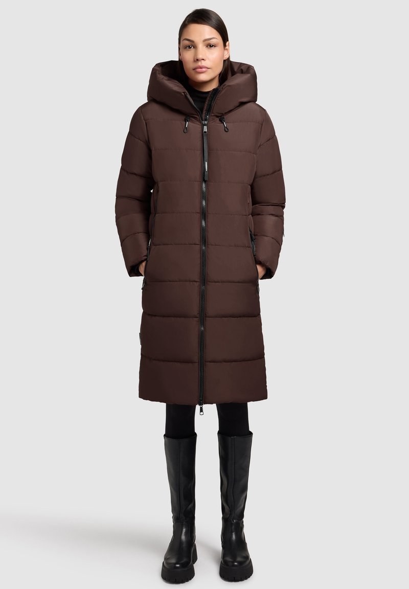 Manteau rembourré brun avec capuche, doté d'une fermeture éclair, de poches latérales et de panneaux matelassés. Porté par-dessus des leggings noirs et des bottes hautes.