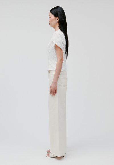 Blusa bianca a maniche corte con texture, abbinata a pantaloni larghi a vita alta color crema, completata da sandali open-toe per un look raffinato.