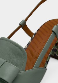 Πράσινο δερμάτινο σανδάλι με φαρδιά λουράκι και λεπτομέρεια φιόγκου· διαθέτει ενσωματωμένη ανάγλυφη σόλα με σχέδιο και καφέ υφασμάτινη σόλα με ανάγλυφα λογότυπα.