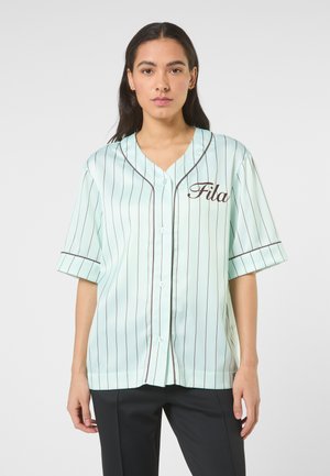 Femme portant une chemise de baseball bleu clair à manches courtes, boutonnée, avec de fines rayures verticales et le logo "Fila" sur la poitrine, associée à un pantalon noir.