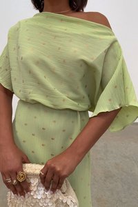 Zelena haljina s teksturom i zlatnim točkicama, off-shoulder kroj i lepršave rukave. Drži bijelu clutch torbicu s perlicama.