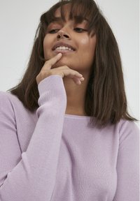 Pulover ribbed de culoare violet deschis, cu un guler larg și mâneci ajustate, având o textură moale și un luciu subtil. Modelul stă cu mâna la bărbie.