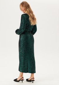 Vestito verde con paillettes, maniche lunghe, vita aderente e gonna lunga fino al pavimento. Presenta un design dritto e una superficie testurizzata. Scarpe con tacco nere.