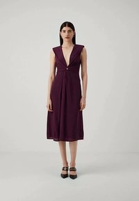 Ženska z dolgimi temnimi lasmi, oblečena v brezrokavni midi obleki globoko vijolične barve z V-izrezom in sprednjim drapiranjem, ki jo dopolnjujejo črne pointed petke.