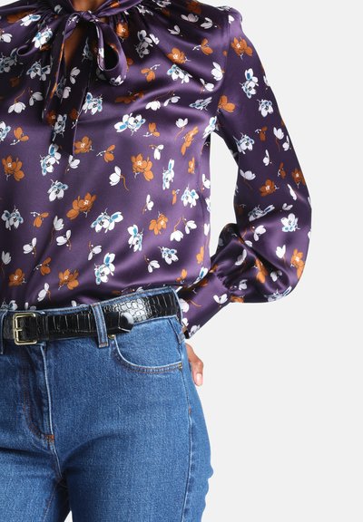 Femme portant un chemisier en satin violet à fleurs avec un col noeud, rentré dans un jean bleu taille haute avec une ceinture noire texturée.