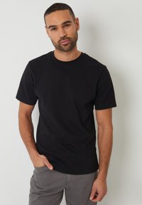 Camiseta negra de cuello redondo con mangas cortas, hecha de una tela suave. El modelo tiene las manos en los bolsillos y lleva pantalones grises.