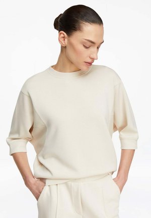 Donna con capelli scuri raccolti in uno chignon, che indossa una felpa cremosa e morbida con maniche lunghe fino al gomito e pantaloni cremisi abbinati, con le mani nelle tasche, che guarda verso il basso.