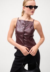 Burgundska umetna usnjena bluza z tankimi naramnicami in sprednjim zapiranjem na gumb. Model je kombiniral z črnimi hlačami in ročno torbico, ter očali v obliki mačjih oči.