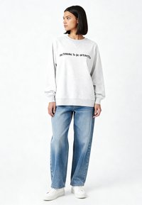 Sweat-shirt gris clair avec "FAIRE SEMBLANT D'ÊTRE INTÉRESSÉ" en texte noir, associé à un jean large bleu clair et des baskets blanches.