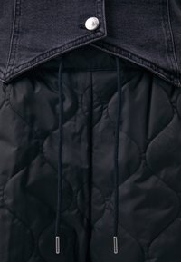 Černá džínová bunda se stříbrným knoflíkem na límci, spárovaná s prošívaným černým materiálem s vlnitým vzorem a šňůrkami.