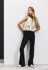 Ärmelloses cremefarbenes Oberteil mit schwarzer Umrandung und Taschen, vorne mit Knöpfen. Kombiniert mit schwarzen, ausgestellten Hosen und beigefarbenen Loafers. Schlichtes Design.