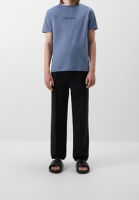 T-shirt in cotone azzurro chiaro con logo ricamato, abbinata a pantaloni neri e sandali neri. Vestibilità semplice e rilassata.