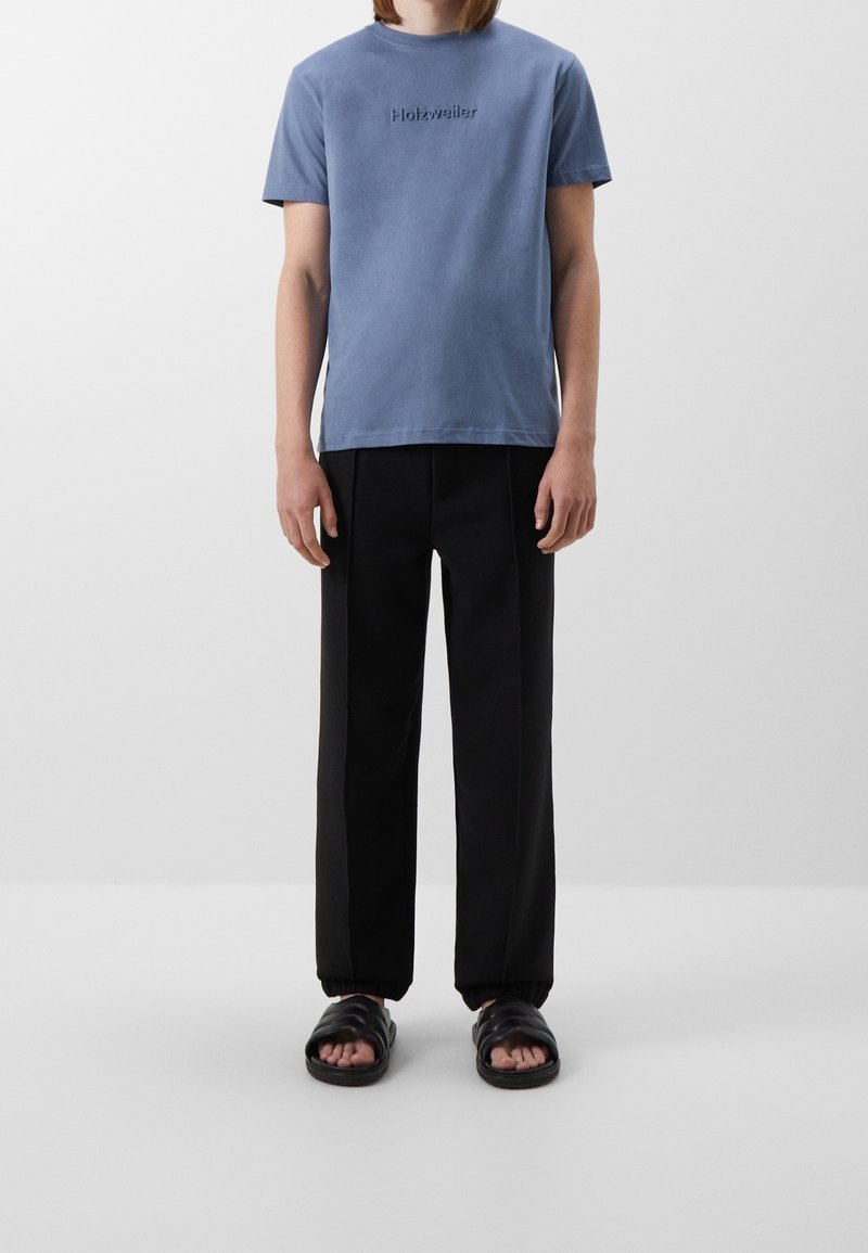 T-shirt in cotone azzurro chiaro con logo ricamato, abbinata a pantaloni neri e sandali neri. Vestibilità semplice e rilassata.