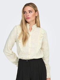 Crèmewitte blouse met bloemrijke ajourmotief, ruches aan de voorkant en knoopsluiting. Lange mouwen met geruchte manchetten. Gecombineerd met zwarte broek.