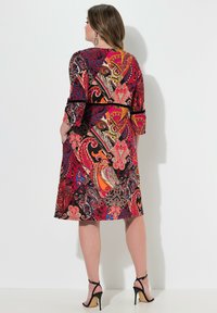 Multicolorkleid mit Paisleymuster in Rottönen, Blau und Orange, mit Dreiviertelärmeln und einem schwarzen Gürtel in der Taille.