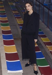 Svart dubbelknäppt blazer med fransdetalj, kombinerad med vida byxor. Bärs med en svart topp och svarta klackar, framför färgglada läktarstolar.