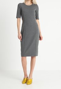 Robe grise jusqu'au genou, manches courtes, coupe ajustée, détails de coutures, texture lisse ; associée à des chaussures jaunes à bout pointu.