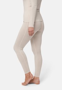 Lähikuva henkilöstä, joka pukeutunut beigeen pitkähihaiseen yläosaan ja vastaaviin legginsseihin, seisoo paljain jaloin yhdellä jalalla hieman kohotettuna.