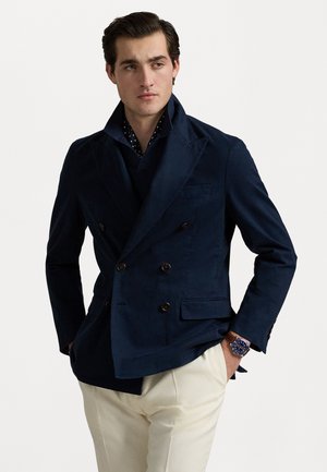Blazer bleu marine à double boutonnage avec de grands boutons, revers pointus et poches latérales, associé à une chemise à pois et un pantalon de couleur claire.