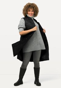 Schwarze gestepptes Weste mit Knopfverschluss, getragen über einem grauen Pullover, kombiniert mit grauen Hosen und schwarzen kniehohen Stiefeln. Glatte Textur, langes Design.