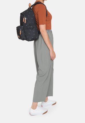 Persoon draagt een roestkleurig kort mouw shirt, grijze broek met wijd uitlopende pijpen, witte sneakers en een zwarte rugzak met een klein licht patroon.