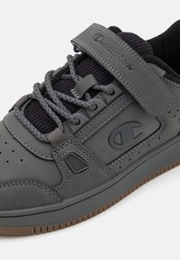 Sneaker Champion di colore grigio scuro con linguetta in maglia nera, lacci grigi, fascia in Velcro e suola in gomma marrone, mostrato in primo piano.