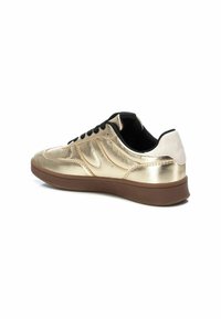 Zapatilla metálica dorada con cordones negros, upper texturizado y contrasta con la pestaña del talón blanca. Cuenta con una suela de goma marrón y forma redondeada.