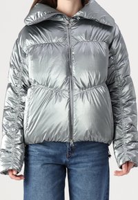 Silver puffer jacket featuring a shiny, metallic finish, oversized collar, and gathered detailing along sleeves and sides. Zippered front closure.