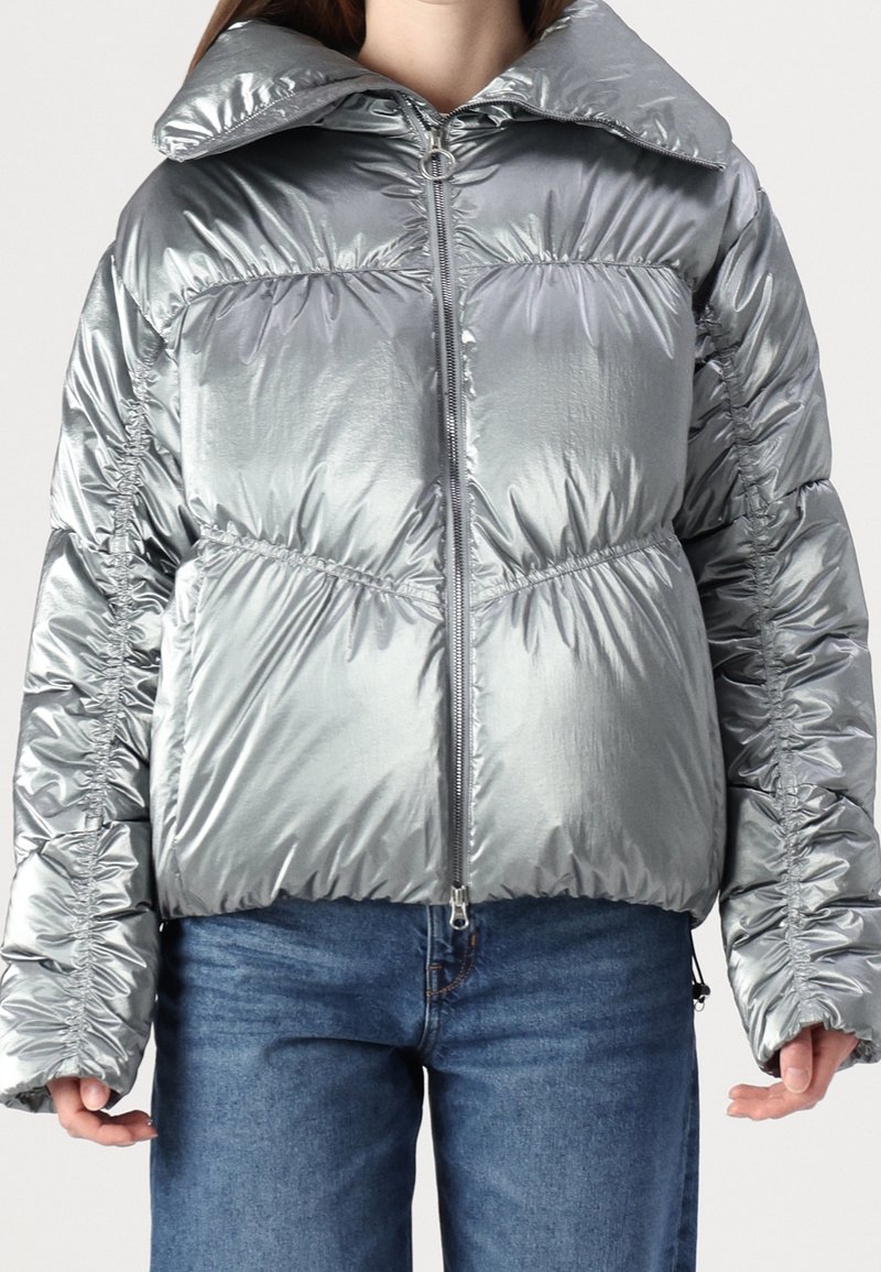 Silver puffer jacket featuring a shiny, metallic finish, oversized collar, and gathered detailing along sleeves and sides. Zippered front closure.