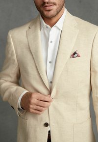 Blazer en lin beige avec un revers cranté et deux boutons, porté sur une chemise blanche. Il est orné d'un pochette de costume à motifs rose et noir.