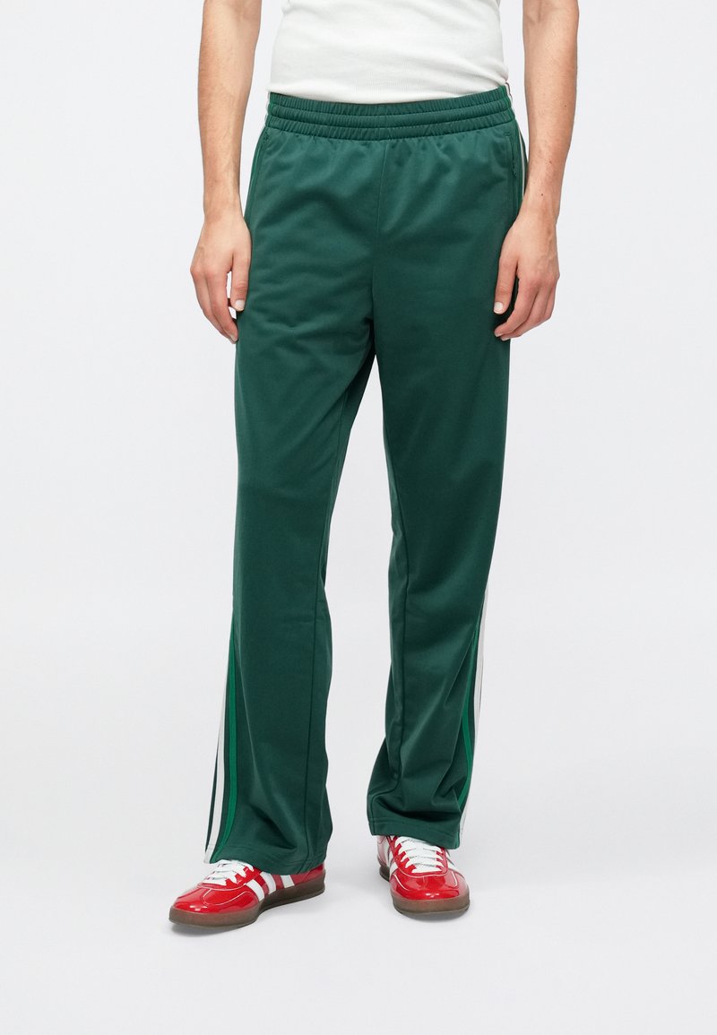 Pantalon de sport en polyester vert avec une ceinture élastique, doté de bandes blanches sur les côtés et une coupe décontractée. Associé à des baskets rouges.