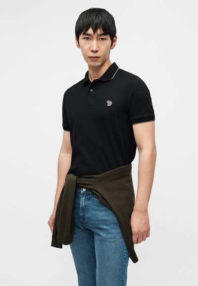 Μαύρο πόλο μπλουζάκι με αντίθετο πράσινο γιακά, διακοσμημένο με μικρό κεντητό λογότυπο. Το μοντέλο φορά μπλε τζιν με καφέ πουλόβερ δεμένο γύρω από τη μέση.