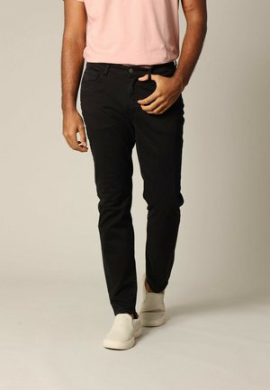 Zwarte slim-fit broeken van katoen, met een riemlus en knoopsluiting, gecombineerd met beige instap schoenen. Model staat tegen een neutrale achtergrond.