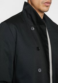 Cappotto nero con colletto alto, dotato di chiusura a doppiopetto, texture liscia e un sottile accento grigio lungo l'imbottitura interna.