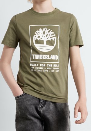Barn iført en olivengrøn Timberland t-shirt med hvidt trælogo og tekst, parret med mørkegrå denimjeans.