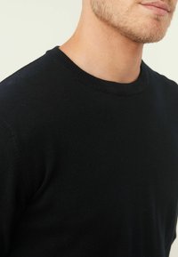 Pull noir à col rond avec une texture lisse, un col ribbed et une coupe ajustée, montré depuis la zone de l'épaule et du cou.