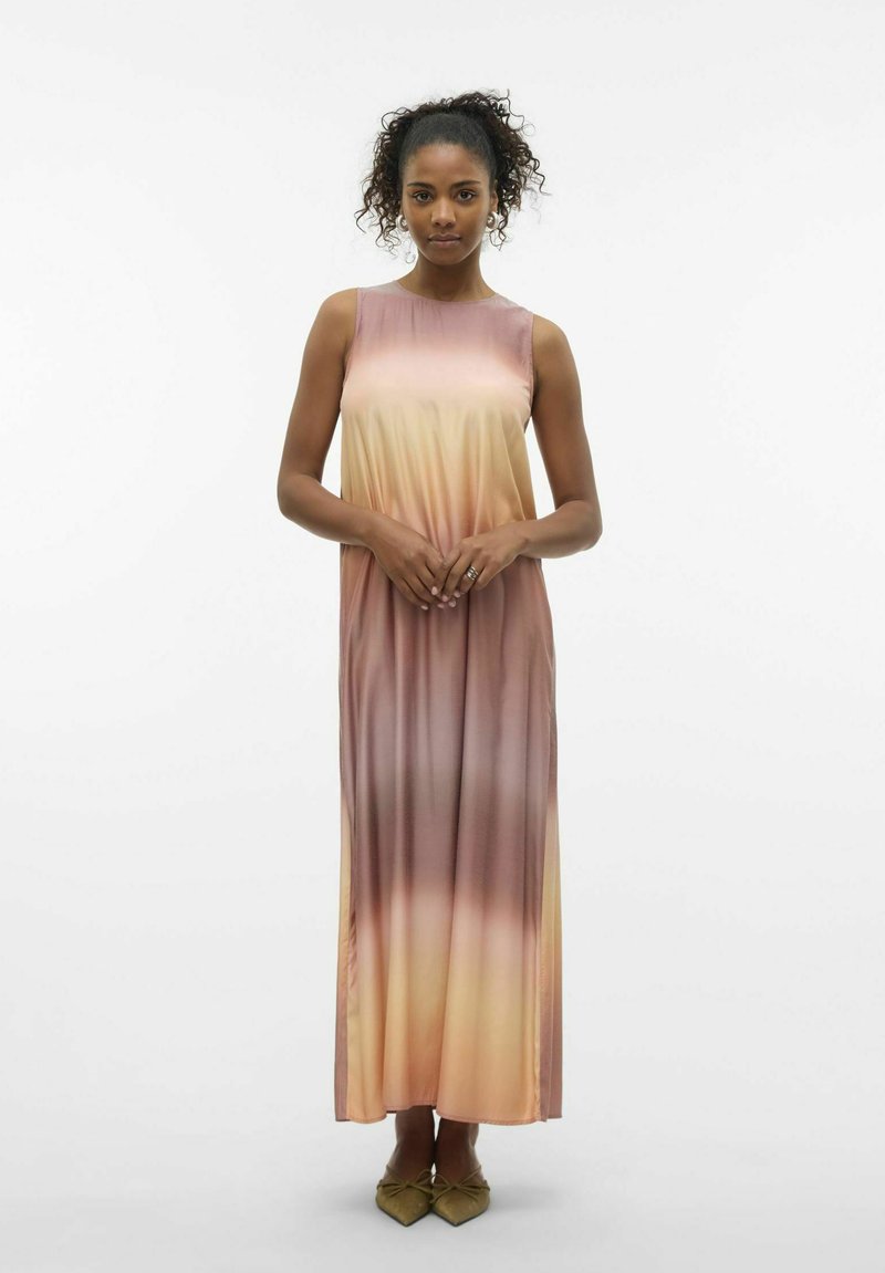 Femme aux cheveux bouclés portant une robe maxi sans manches dégradée rose et beige, debout les mains jointes devant un fond blanc uni.