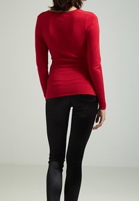 Donna che indossa una maglietta rossa a maniche lunghe aderente e pantaloni neri, in piedi con le spalle rivolte verso la macchina fotografica su uno sfondo neutro.