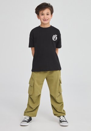 T-shirt nera in cotone con logo bianco sul petto, abbinata a pantaloni cargo verde oliva con tasche laterali e orli alla caviglia, indossati con sneakers nere.
