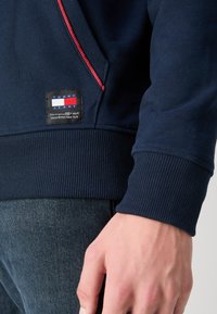 Tmavomodrý sveter s rebrovanými manžetami, červeným zipsom a nášivkou s textom "TOMMY JEANS" na prednej strane. Mäkko bavlnené materiály.