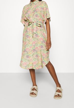 Knielanges Kleid mit floralem Muster in Hellgrün, mit Kragen, kurzen Ärmeln, tailliertem Gürtel und leichtem Stoff. Kombiniert mit beigen Sandalen.